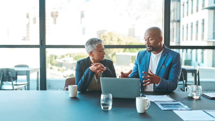 Two business people sit at a conference table with a laptop and papers in discussion.