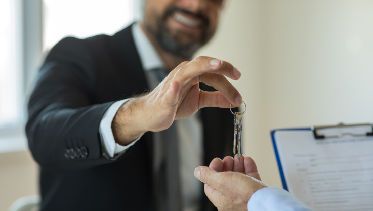 A man extends his hand over a desk in an office holding a house key over the hand of a customer. 