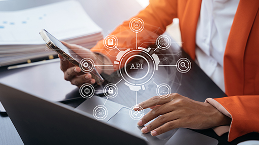 A woman in an orange blazer sits at a laptop with her mobile phone in hand. A technology style graphic with the letters API are across the top of the image.