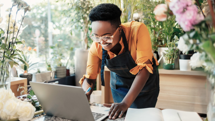 The owner of a flower shop stands at a table working on their laptop. 