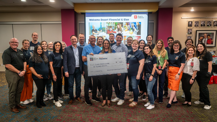 A large group of volunteers stand together smiling at the camera with those in the center holding a large check. 